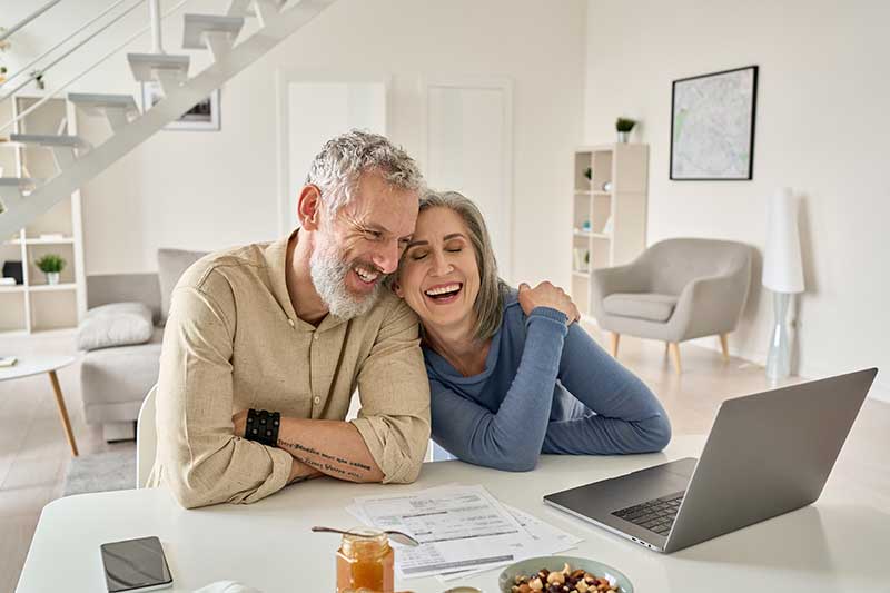 probate administration attorney massachusetts - Photo description: Smiling middle aged senior 50s husband and wife looking at laptop, at home, in virtual meeting with attorneys, perhaps. 